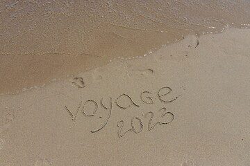The inscription voyaje 2022 in French, travel 2022, on the sand by the water and the rising wave, seashore beach vacation by the sea