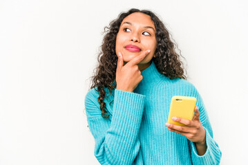 Young hispanic woman holding mobile phone isolated on white background looking sideways with doubtful and skeptical expression.