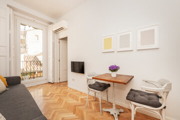 Living room with wooden table with cast iron foot, white wooden chairs, balcony with black metal railing and views of the city's rooftops