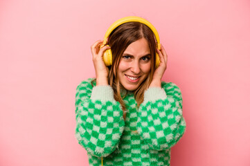 Young caucasian woman listening to music isolated on pink background covering ears with hands.
