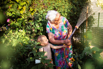 Cute blond toddler boy helping grandmother in the garden. Grandson watering plants garden vwith water hose in backyard at summer. Generation concept.