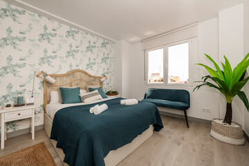 Bedroom with a double bed with a wicker headboard, walls with decorative wallpaper with plant motifs, a natural fiber rug, a blue blanket and a matching velvet armchair