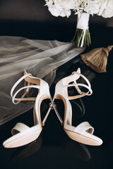 Wedding. Details. The bride's white shoes lie on a black table along with rings, next to it is a bouquet of white flowers with a silk ribbon.