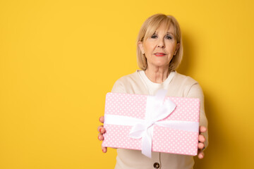 Portrait of cheerful positive glad charming aged woman holding gift box in pink package standing isolated over yellow background.