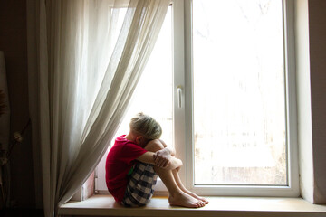 Sad teenager boy sitting on the sill of the large window in a dark room and looks forward.