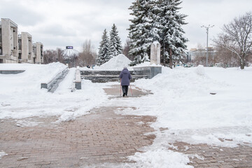 An elderly woman with canes in her hands walks along a snow-covered path in the city. An elderly woman trains Nordic walking with sticks in the city in spring in April.