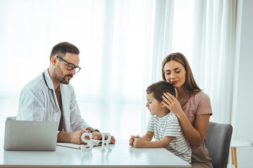 Smiling male pediatrician in white medical uniform holding clipboard, listening to young mother with kid son at checkup meeting, professional children doctor consultation, healthcare concept.