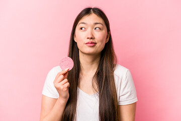 Young asian woman holding facial sponge isolated on pink background dreaming of achieving goals and purposes
