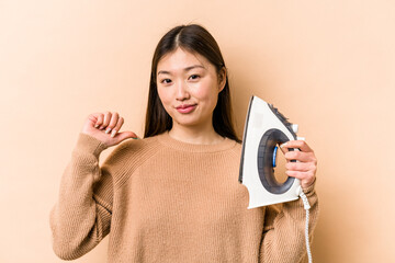 Young asian woman holding iron isolated on beige background feels proud and self confident, example...