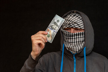 A man in a hoodie examines a $100 bill, his face is covered with an Arab scarf, close-up, selective...