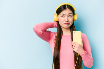 Young asian woman listening to music isolated on blue background touching back of head, thinking...