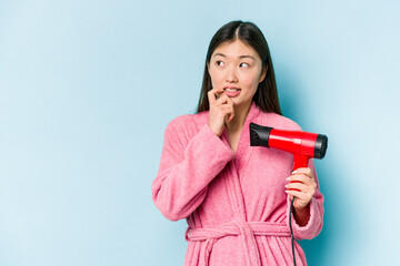 Young asian woman wearing a bathrobe and holding hairdryer isolated on pink background relaxed thinking about something looking at a copy space.