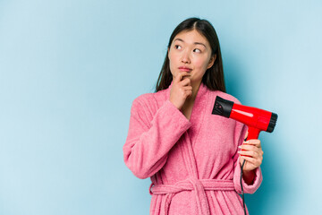 Young asian woman wearing a bathrobe and holding hairdryer isolated on pink background looking sideways with doubtful and skeptical expression.