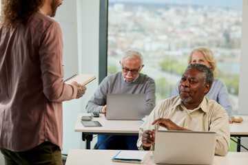 A group of multicultural senior people is listening to a lesson and typing on their laptops in the classroom.
