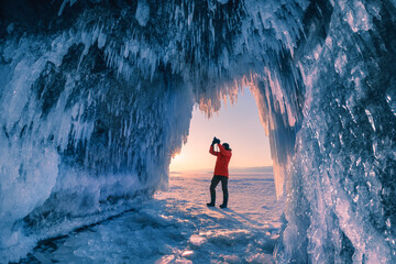 Man tourist photograph the ice cave on Baikal lake at sunset. Winter landscape of Baikal lake,...