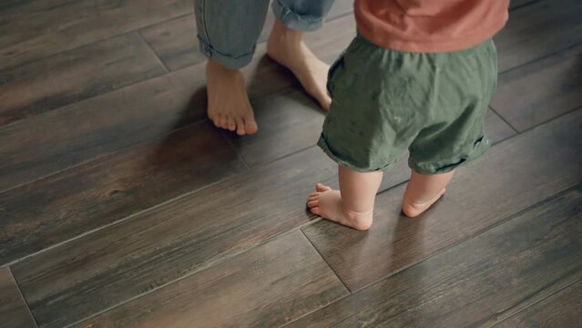 Little feet walking on floor, close-up. Baby learning to walk at home. Baby first steps. Smiling caring mother and cute little infant child girl cuddling in bedroom. Mum and child tender moments.