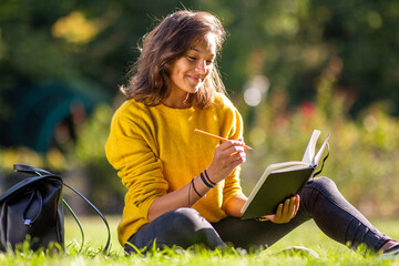 smiling young woman sitting on grass in park writing in book