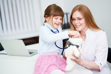 Caucasian female doctor wear face mask show little girl examining bear toy by stethoscope indoor hospital