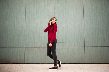Full body young woman walking and talking with cellphone by wall