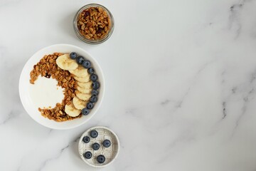 Bowl with granola, yougurt, banana and blueberry, jar with granola, saucer with blueberry, marble background, flat lay