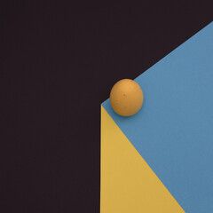 A yellow Easter egg with a sharp shadow stands on the edge of a blue-yellow cube on a black background. Minimal Easter scene.