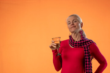 Beautiful smiling happy blond mature woman holding wineglass and drinking wine on solid orange background with copy space sitting on stool.