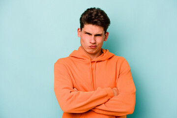 Young caucasian man isolated on blue background frowning face in displeasure, keeps arms folded.