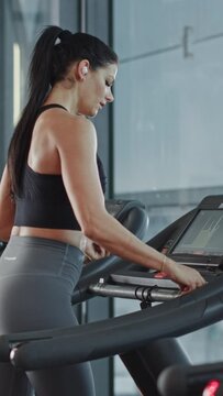 Beautiful Athletic Sports Woman in the Gym Puts on Wireless Headphones, Turns on Podcast or Sport Music Playlist with Smartphone and Starts Running on Treadmill. Vertical Screen Orientation Video 9:16