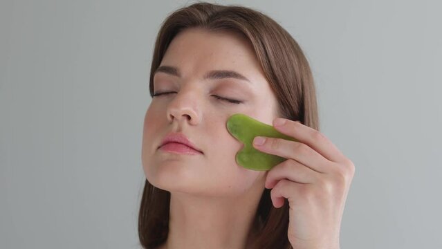 Beautiful naked girl with closed eyes using facial gua sha jade board isolated on beige.