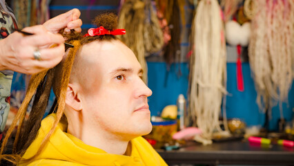 Unrecognizable person makes hairstyle for young man in salon. Professional hairstylist makes brown dreadlocks for client.
