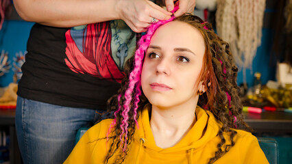 Unrecognizable person makes curly hairstyle for young woman in salon. Professional hairstylist makes pink and brown dreadlocks for client.