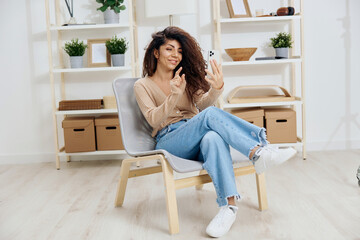 Enjoyed happy concentrated tanned curly Latin lady have video call with trendy smartphone sit on chair in home modern interior. Copy space Mockup Banner. Concept distance communication