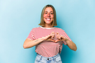 Fototapeta na wymiar Young caucasian woman isolated on blue background smiling and showing a heart shape with hands.