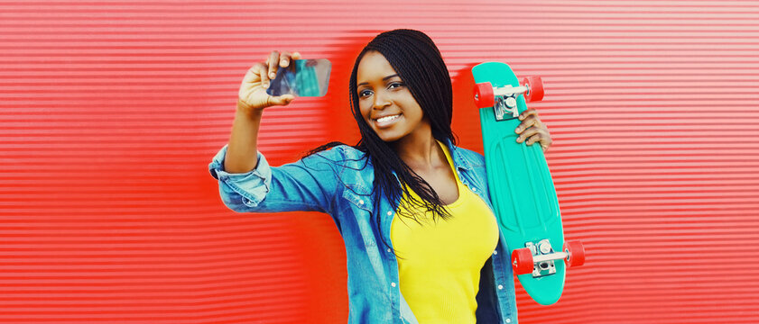 Summer portrait of happy smiling young african woman taking selfie by smartphone with skateboard on vivid background