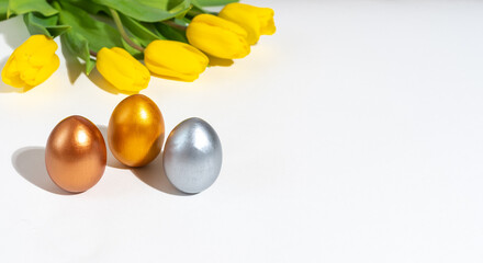 Three multi-colored painted Easter eggs on a table next to yellow tulips on a white background. Spring holiday
