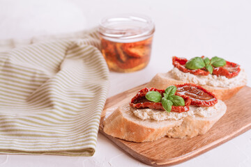 Bruschetta or toast with ricotta cheese and sliced sun dried tomatoes topped with basil on wooden cutting board, white table background. Selective focus