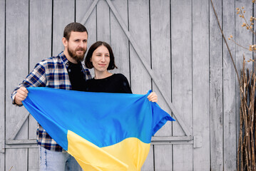 Young family, husband and wife hold blue and yellow state flag of Ukraine on gray wooden wall on street outside in snowy weather. Support for the country during the occupation.