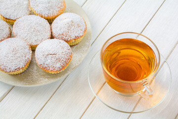 Freshly baked appetizing cupcakes sprinkled with powdered sugar on a plate and a cup of tea in a transparent cup on a white wooden table