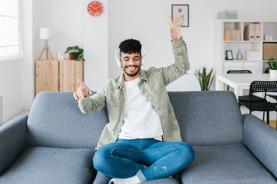 Young joyful man wearing headphones listening music and dancing while sitting on sofa at home indoors on weekends free time. People lounge lifestyle concept