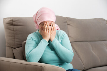 Sad woman with cancer in head scarf crying desperately alone sitting on a couch at home in a dark winter day