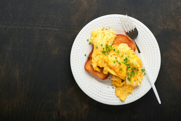 Scrambled Eggs. Fluffy and Buttery scrambled eggs on bread with microgreen radish and hollandaise sauce on white plate over dark old wooden background. Homemade breakfast or brunch meal. Top view.