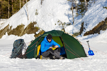 Man looks at the weather forecast in smartphone in green tent with backpack and snow shovel in the...