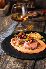 On a serving board, mashed sweet potatoes with bacon rolls with sun-dried tomatoes, microgreens and onion chips. On a wooden table is a box with spices and herbs, a glass of cognac. Restaurant menu