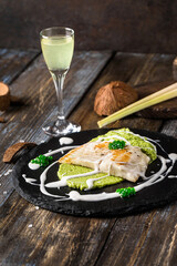 On a round serving board mashed broccoli and vegetables, grilled fish fillet with white cream sauce. On a wooden table a glass of limoncello, spices, lemongrass. Restaurant menu. Close up.