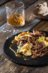 On a round serving board, mashed potatoes with beef in mushroom sauce, with herbs. On a wooden table is spices, mushrooms, a glass of cognac. Close up. For the menu of a restaurant, cafe, bar