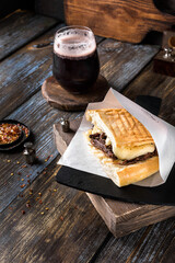 Sandwich with beef, cheese and sauce on ciabatta in a paper cover on a wooden table. A glass of dark beer and spices in the composition. Dish for the menu of a restaurant, bar, cafe. Close up
