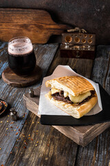 Sandwich with beef, cheese and sauce on ciabatta in a paper cover on a wooden table. A glass of dark beer and spices in the composition. Dish for the menu of a restaurant, bar, cafe. Close up