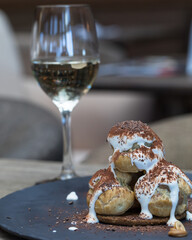 On black plate a slide of profiteroles poured with buttercream and sprinkled with chocolate powder. Next to a glass of white wine. The blurry interior of the restaurant is in the background. Close up.