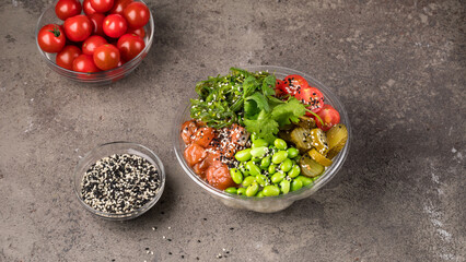 On a gray textured background, a poke bowl with rice, salmon beans edamame chukka, cherry tomatoes pickled cucumbers sesame and parsley.  Bowl of sesame seeds and a bowl of cherry tomatoes. 