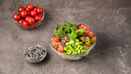 On a gray textured background, a poke bowl with rice, salmon beans edamame chukka, cherry tomatoes pickled cucumbers sesame and parsley.  Bowl of sesame seeds and a bowl of cherry tomatoes. 